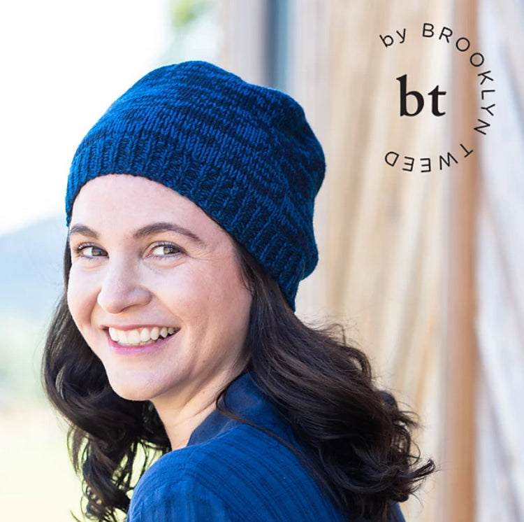 A young white woman with long brown hair smiles, wearing a marled handknit hat. 