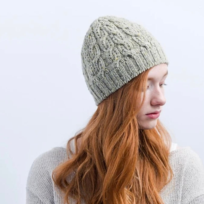 A young white woman with long red hair looks downward, wearing a gray cabled hat. 