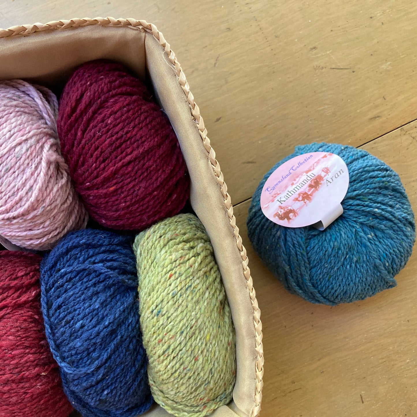 Colorful balls of tweed Queensland Kathmandu Aran yarn in a rectangular basket on a wooden surface. 