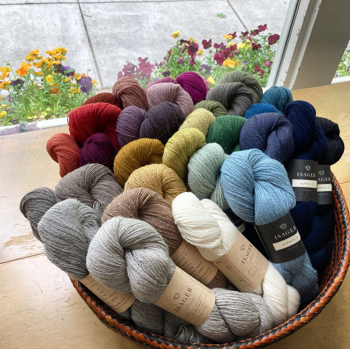 Basket of colorful skeins of Isager Alpaca 2 yarn on a wooden table in front of a window.  