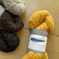 Four skeins of Fibre Co. Arranmore Light yarn in black, brown, beige, and yellow, laying on a wooden surface. 