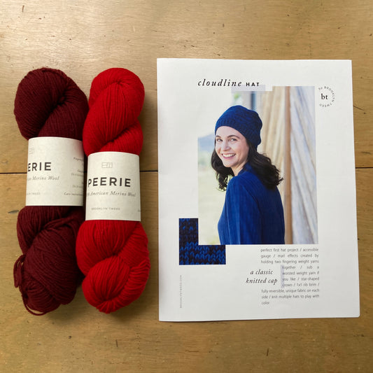 Two skeins of Brooklyn Tweed Peerie yarn and a print copy of a hat pattern lie on a wooden surface. 