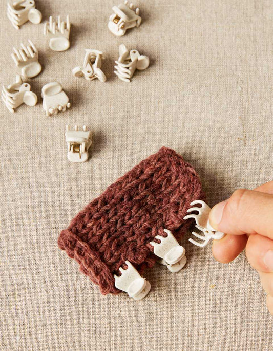 Cocoknits claw clips scattered on a light gray surface, with a rust colored knit swatch. 