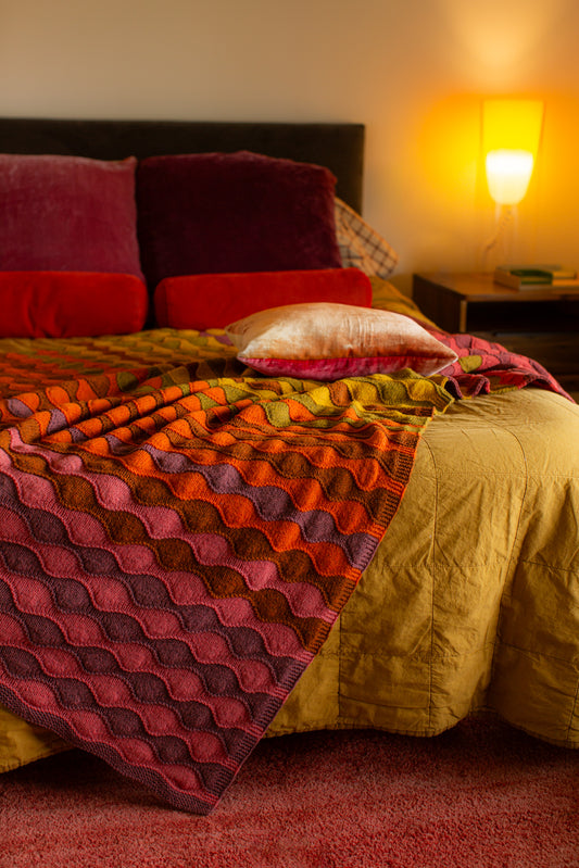 Bed draped with red velvet pillows and warm toned hand knit blanket, from Brooklyn Tweed's Knit & Crochet Blankets book.