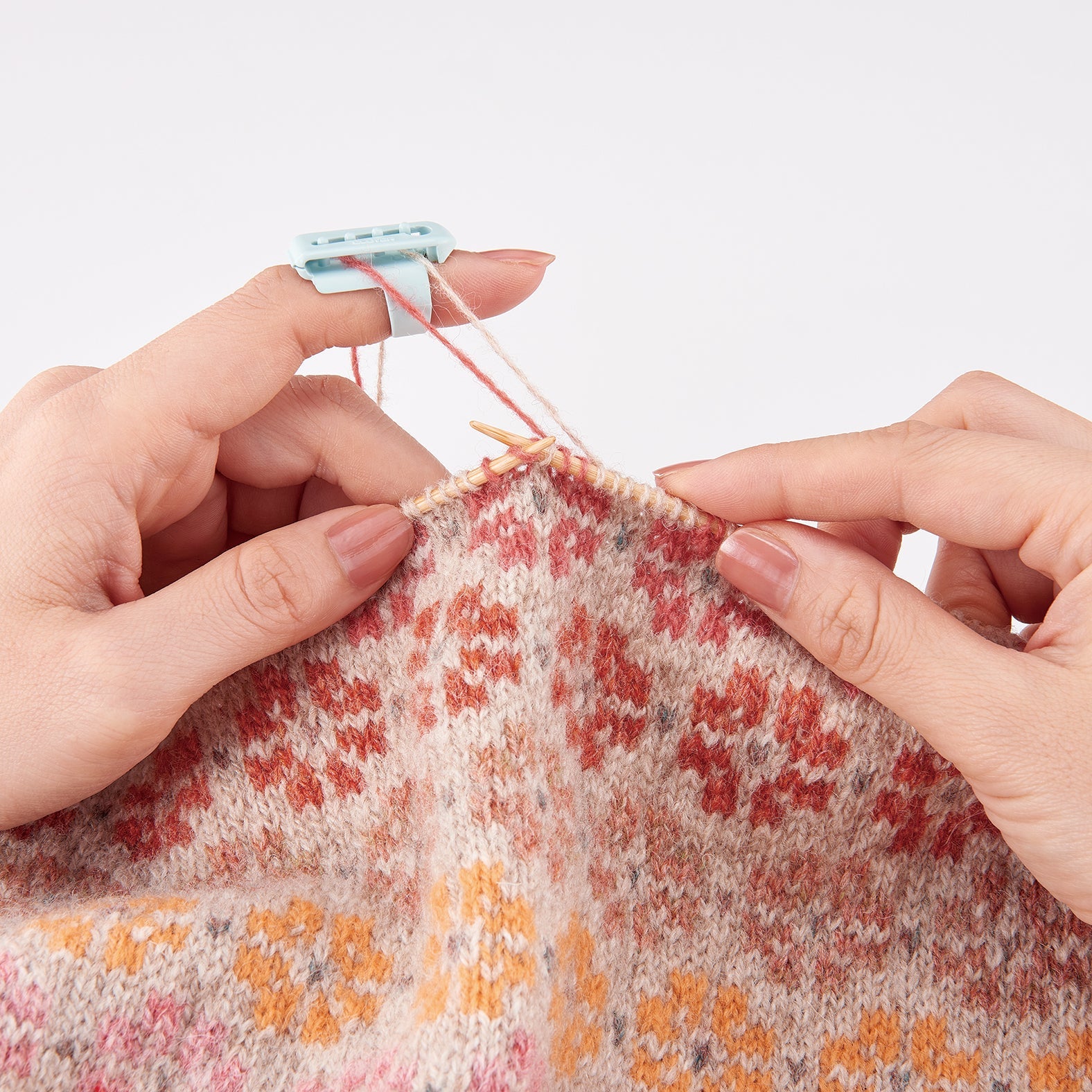 Clover yarn guide attached to knitters' left pointer finger, threading a red strand and a beige strand while knitting a floral  colorwork patern.