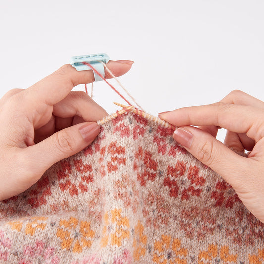 Clover yarn guide attached to knitters' left pointer finger, threading a red strand and a beige strand while knitting a floral  colorwork patern.