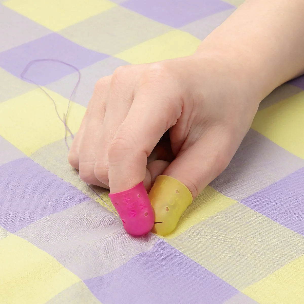 Close-up of a hand wearing the Clover Flexible Thimble on both thumb (large, yellow) and pointer finger (small, pink) while using a sewing needle on checkered fabric.
