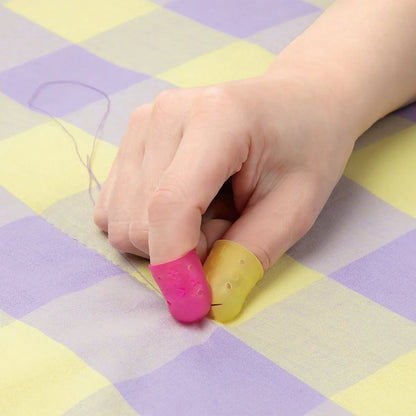 Close-up of a hand wearing the Clover Flexible Thimble on both thumb (large, yellow) and pointer finger (small, pink) while using a sewing needle on checkered fabric.