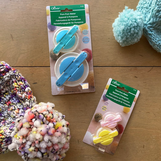 Two packages of pom-pom makers on a wooden surface with colorful pom-poms.