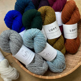 Colorful skeins of Brooklyn Tweed Loft yarn in a round basket on a wooden surface. 