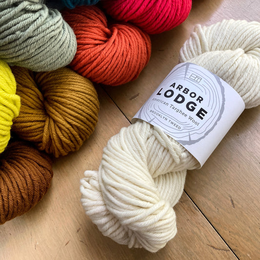 Natural white skein of Brooklyn Tweed Arbor Lodge laying on a wooden surface, near a colorful pile of skeins. 