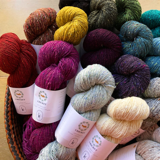 Colorful skeins of Kelbourne Woolens Cricket yarn in a basket on a wooden surface. 