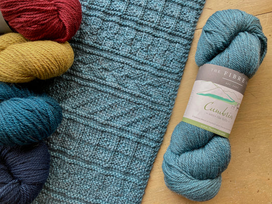 Close up on a textured scarf knit with Fibre Co. Cumbria, laying on a wooden surface near skeins of yarn. 