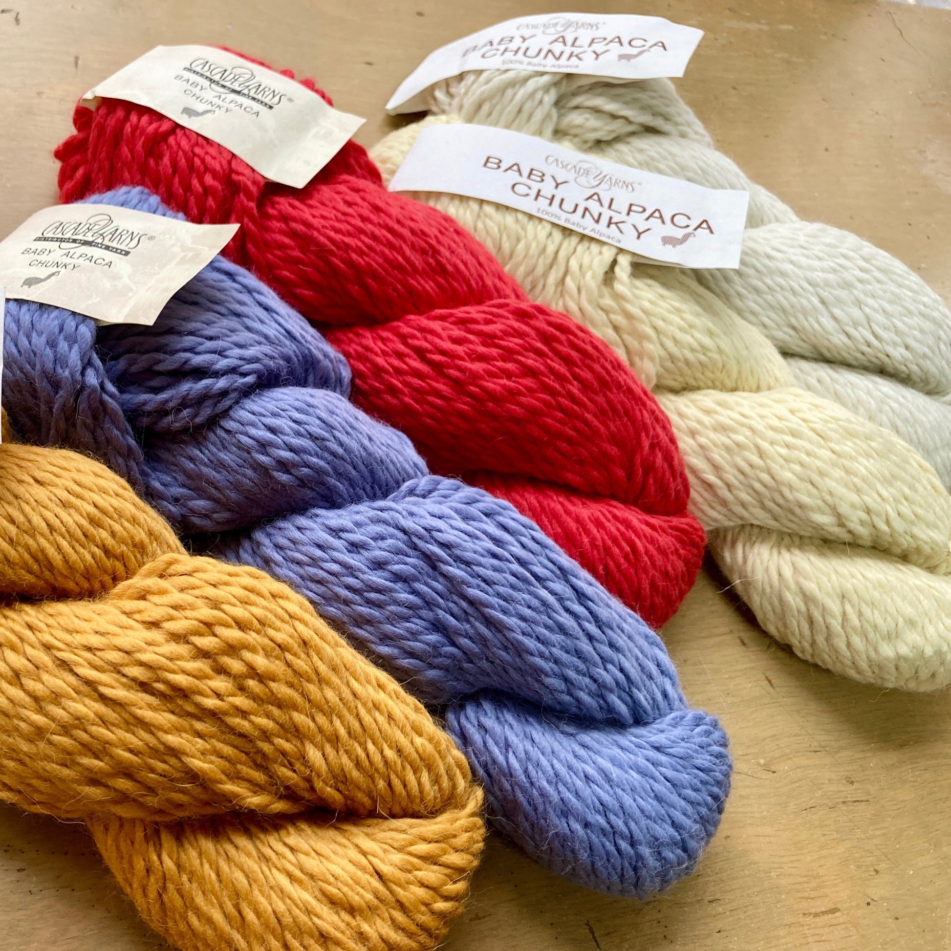 Five colorful skeins of Cascade Baby Alpaca Chunky lying on a wooden surface. 