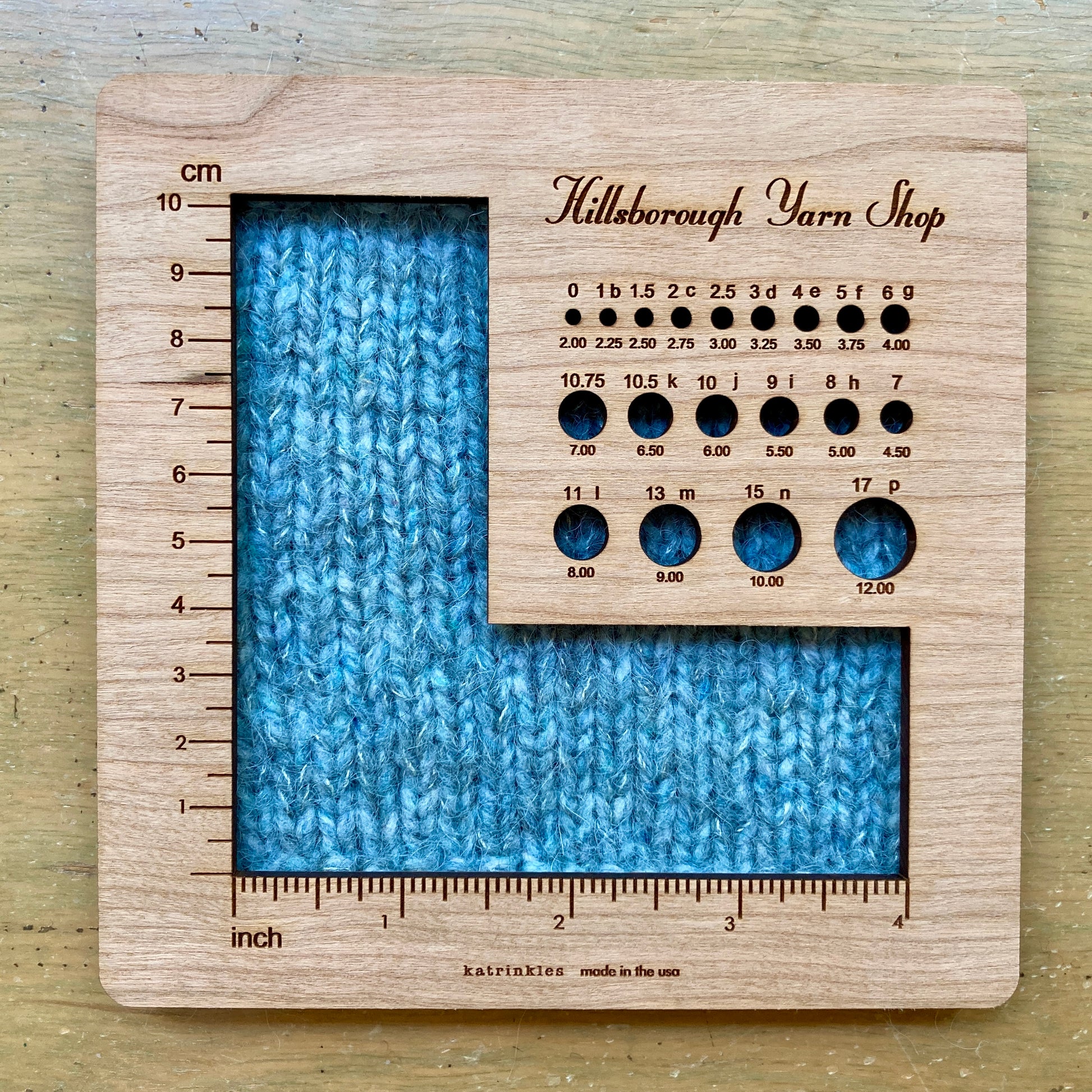 Square wooden Hillsborough Yarn Shop gauge tool with round openings for measuring needles and hooks, and  a larger L shaped opening for each measuring swatches. 