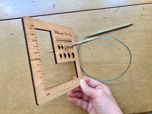 Hand holding a square wooden Hillsborough Yarn Shop gauge tool with round openings for measuring needles and hooks, and a larger L shaped opening for each measuring swatches. A silver circular needle with a blue cord is poking through one of the holes. 