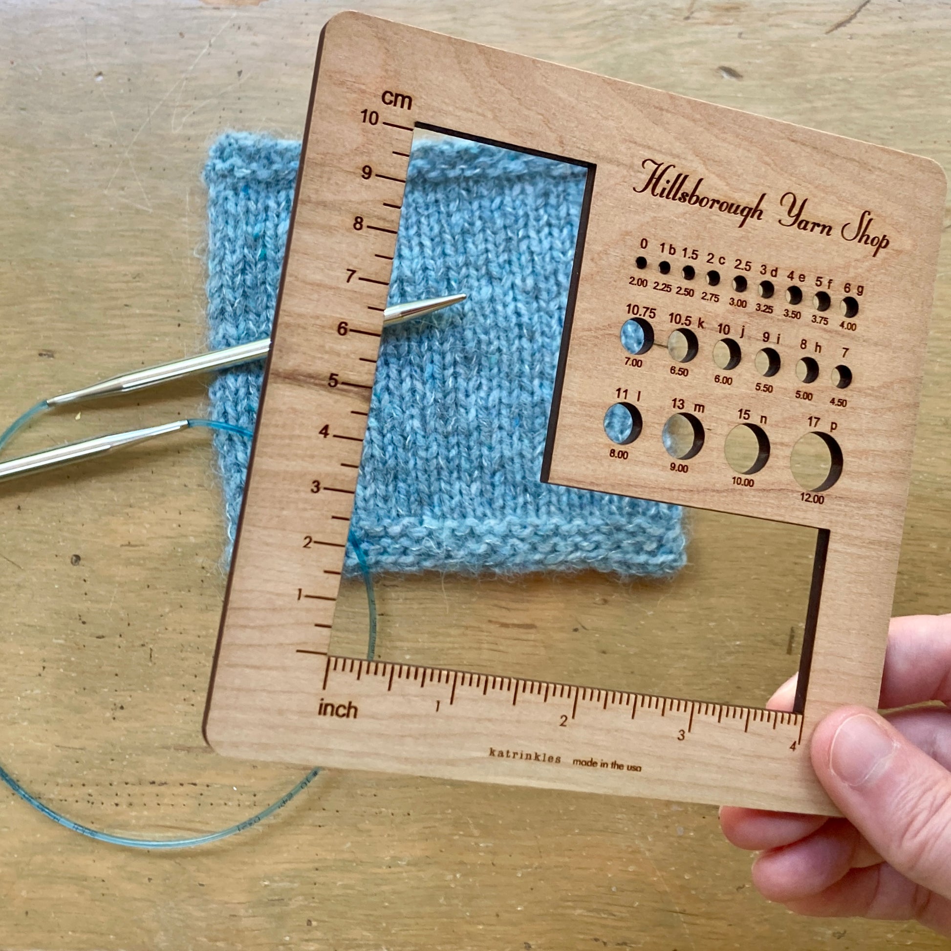 Hand holding a square wooden Hillsborough Yarn Shop gauge tool with round openings for measuring needles and hooks, and a larger L shaped opening for each measuring swatches.  Below, a light blue swatch and pair of silver needles rest on a wooden surface. 