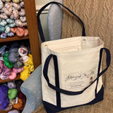 Hillsborough Yarn Shop tote bag on display near shelves of colorful yarn. 