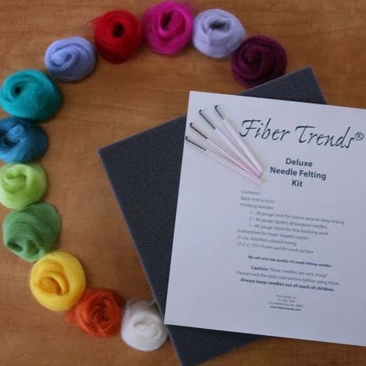 Puffs of colorful roving arranged in a circle on a wooden surface, next to a gray needle felting pad and several needles. 