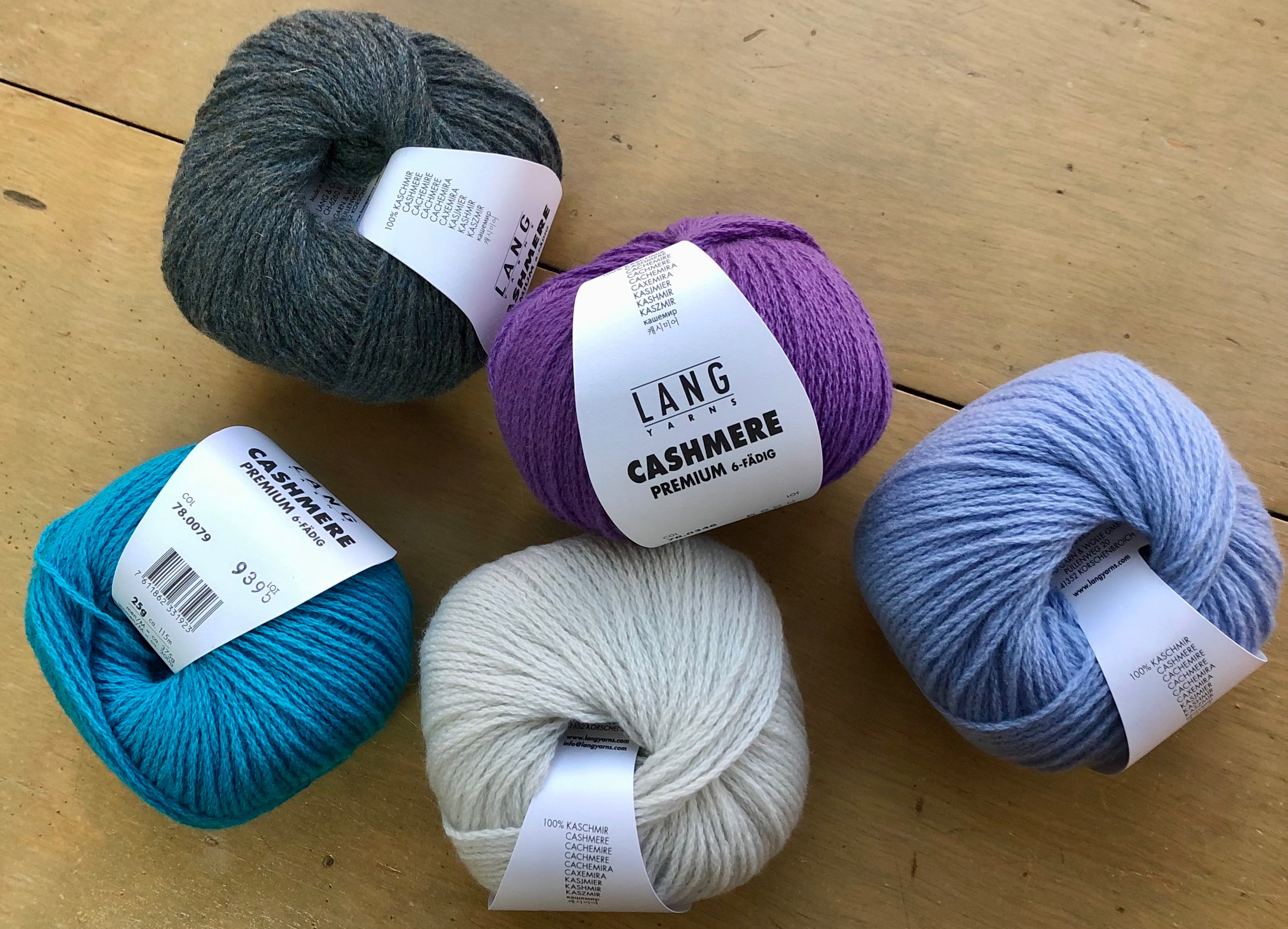Five skeins of yarn in different colors with visible labels on a wooden surface.