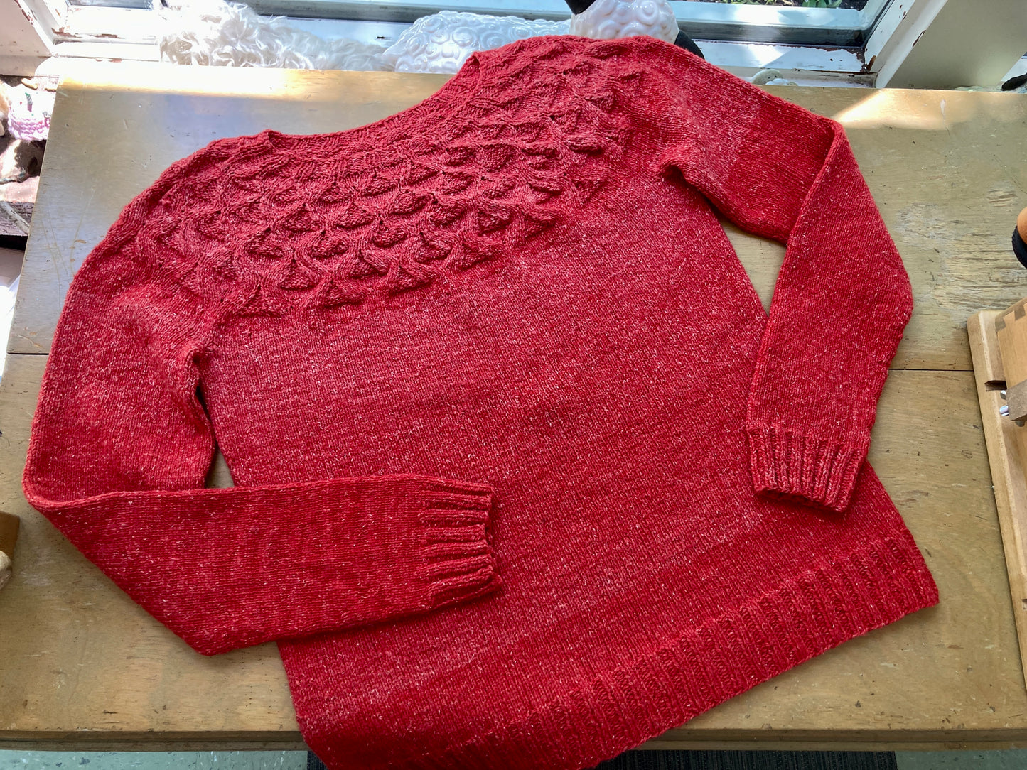 Handknit textured red sweater, lying on a wooden table with a ballwinder attached. 