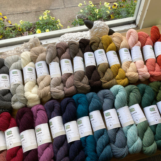 Many colorful skeins of Kelbourne Woolens Camper yarn, arranged on a table in front of a window. 