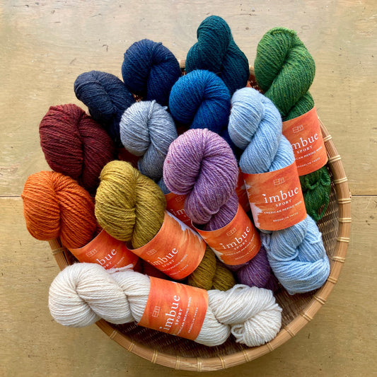 Colorful skeins of Brooklyn Tweed Imbue Sport yarn in a round basket on a wooden surface. 