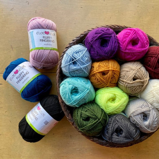 Colorful balls of Ewe Ewe Fluffy Fingering yarn in a round basket on a wooden surface. 