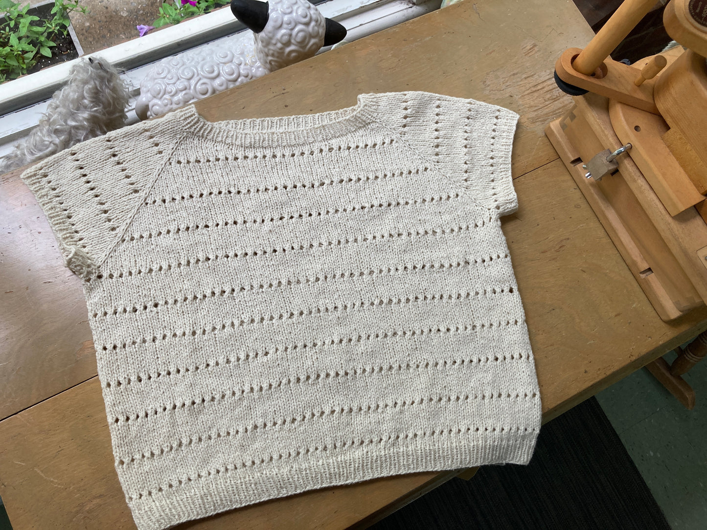 Handknit cream colored eyelet tee, lying on a wooden table with a ballwinder attached. 