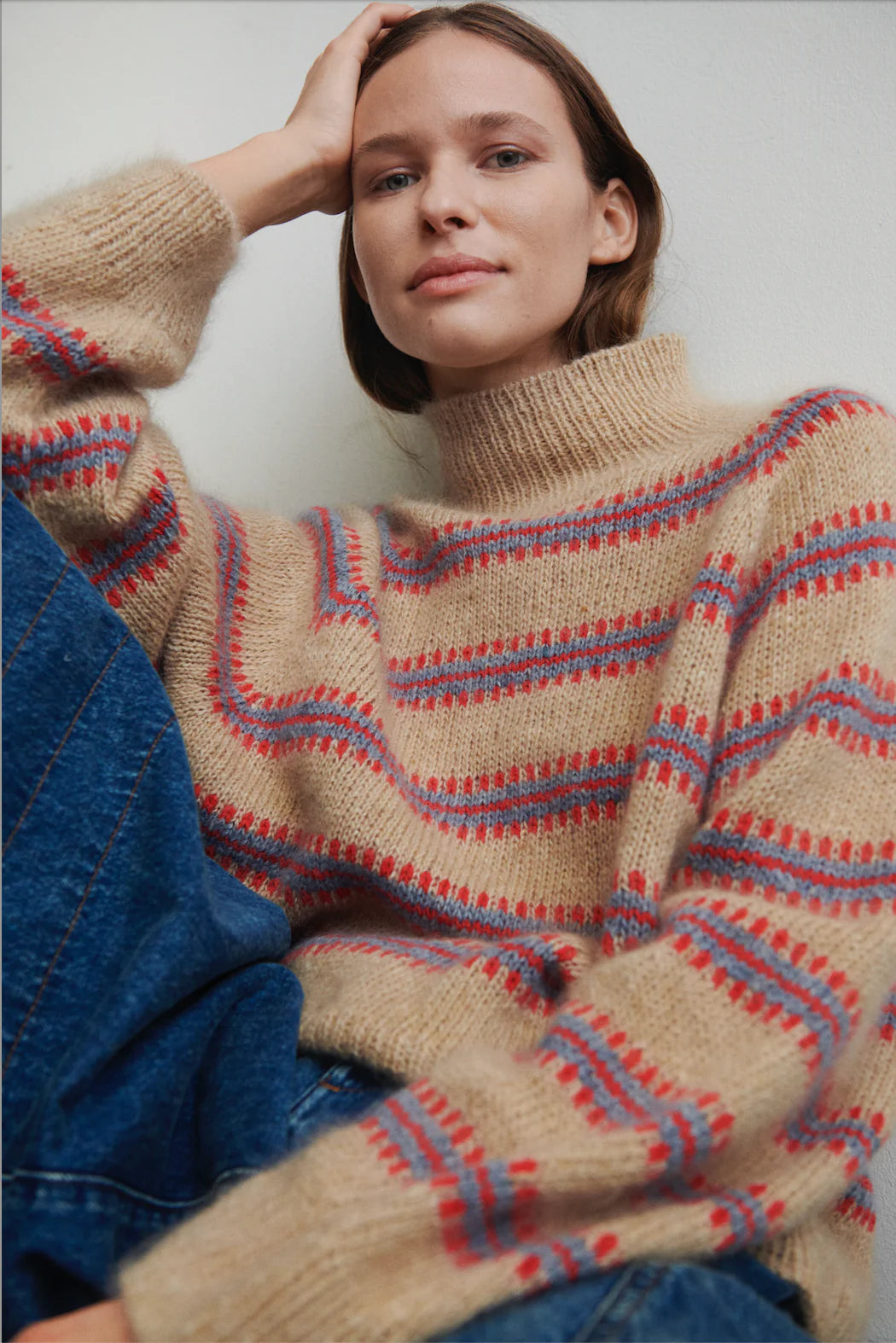 Model sits, propping head on hand against white backdrop wearing the Norma sweater, designed by My Favourite Things Knitwear as part of Isager Archives collection, in sand color with light blue and red stripe motif.