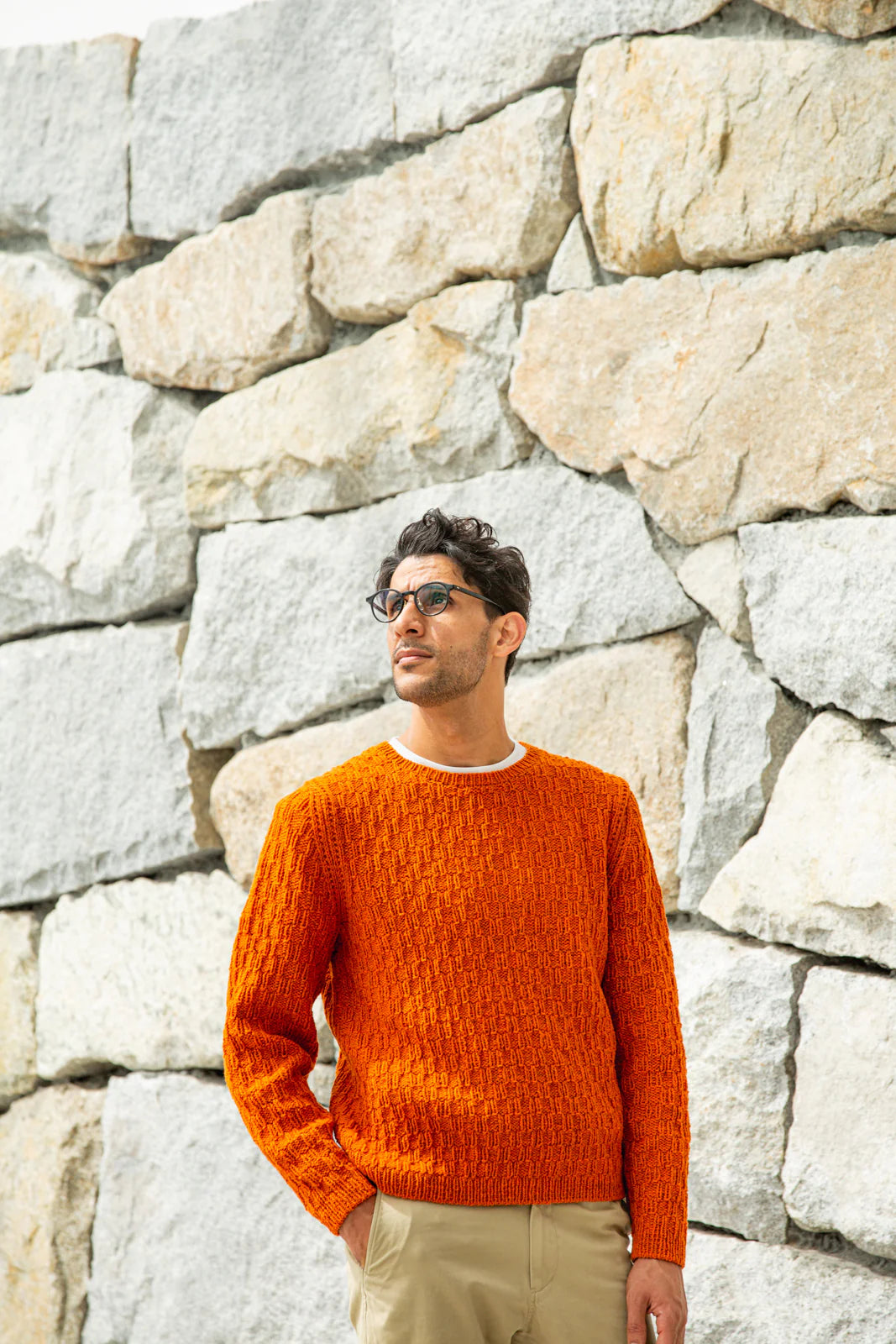 Person wearing orange handknit sweater stands before rock wall, showing off a pattern from Brooklyn Tweed’s Northern Lights book. 