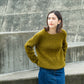 Person wearing moss green handknit sweater stands before concrete wall, showing off a pattern from Brooklyn Tweed’s Northern Lights book. 