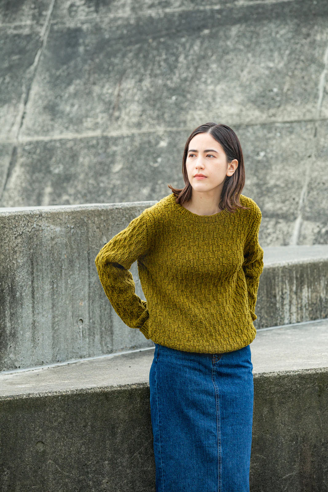 Person wearing moss green handknit sweater stands before concrete wall, showing off a pattern from Brooklyn Tweed’s Northern Lights book. 