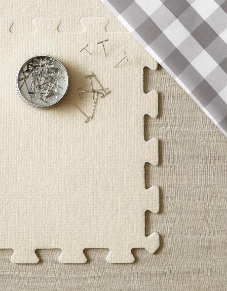 Square gray tile with puzzle piece edges, an open tin of t-pins, and a gray and white gingham cloth arranged on a beige surface. 