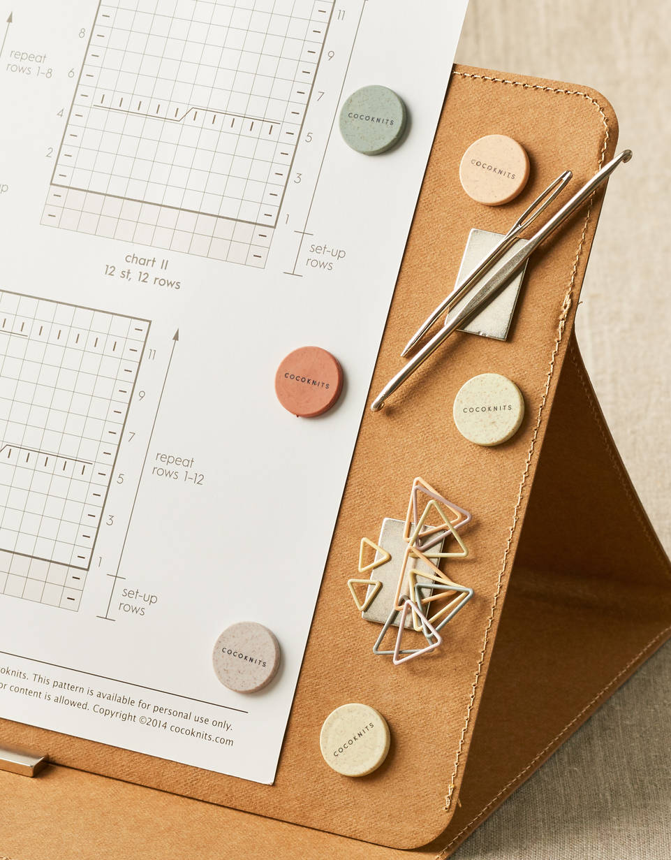 Kraft brown Cocoknits maker’s board covered in magnets which hold up a knitting pattern and various knitting tools. 