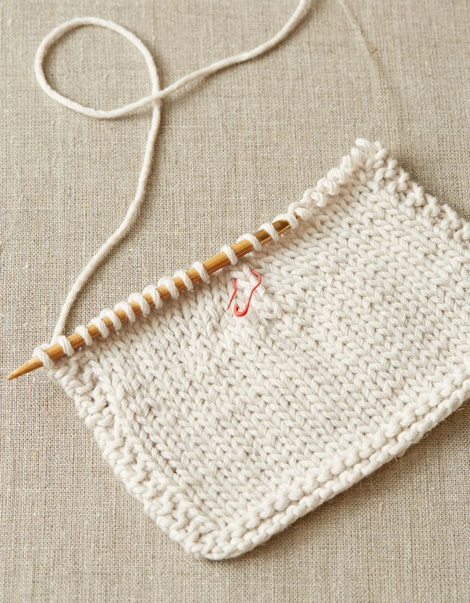Cream colored knit swatch in progress, with a red opening stitch marker near the wooden needle. 