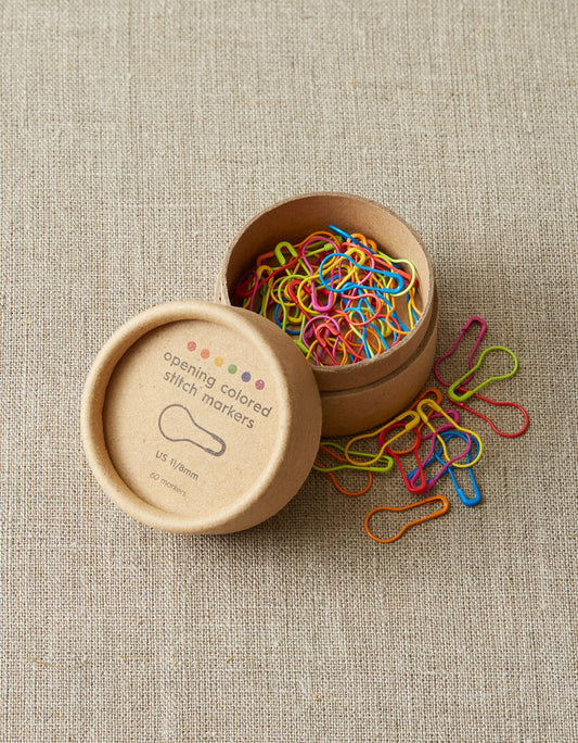 Open box of colorful cocoknits stitch markers on a light gray surface. 