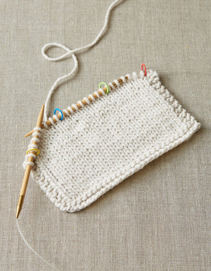 White knit swatch in progress, with colorful markers at four points along the wooden knitting needle. 