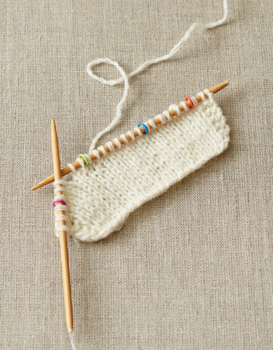 White knit swatch in progress, with colorful markers at four points along the wooden knitting needle. 