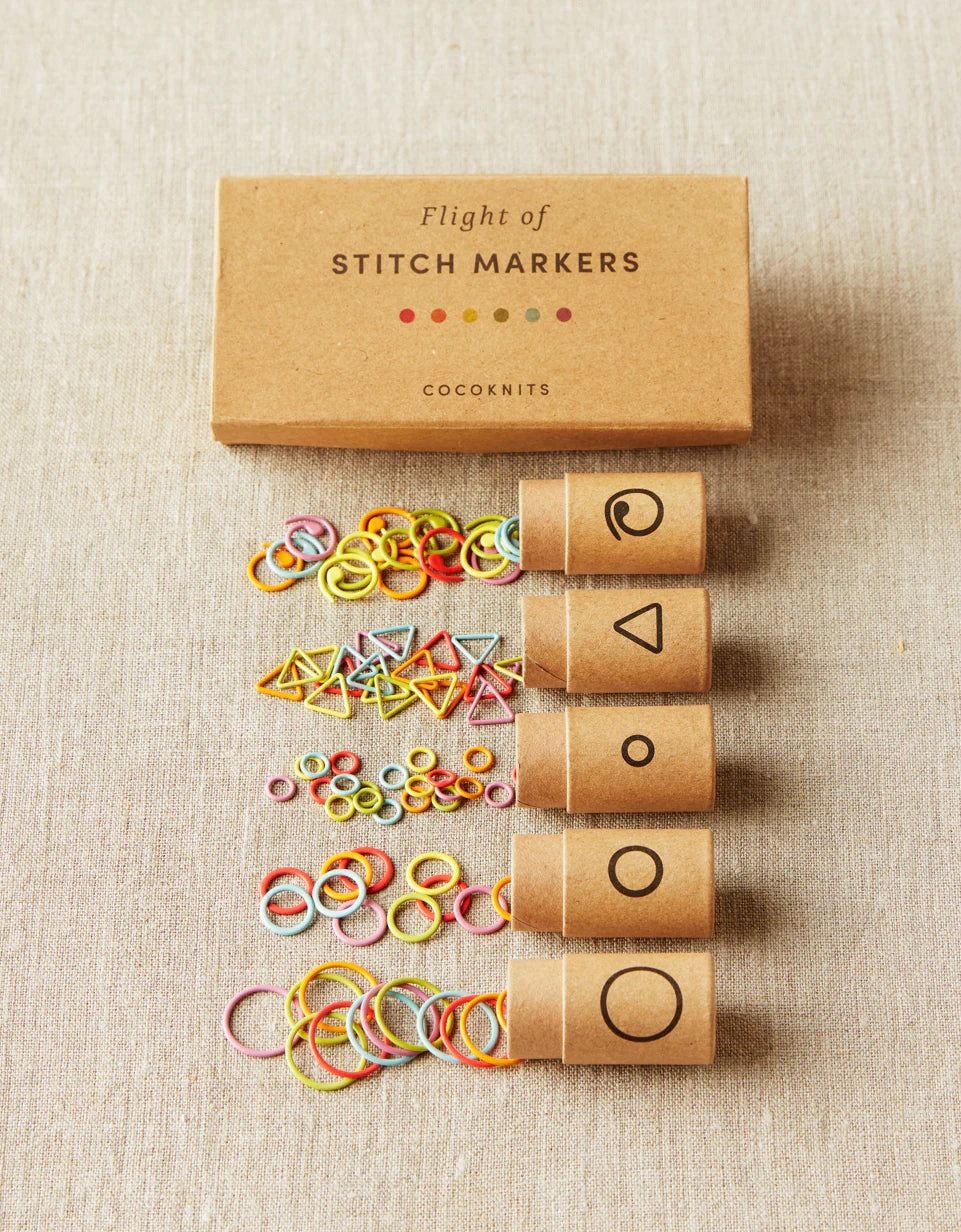 Kraft brown box of cocoknits Flight of Stitch Markers, lying on a gray surface with five open containers of colorful stitch markers. 