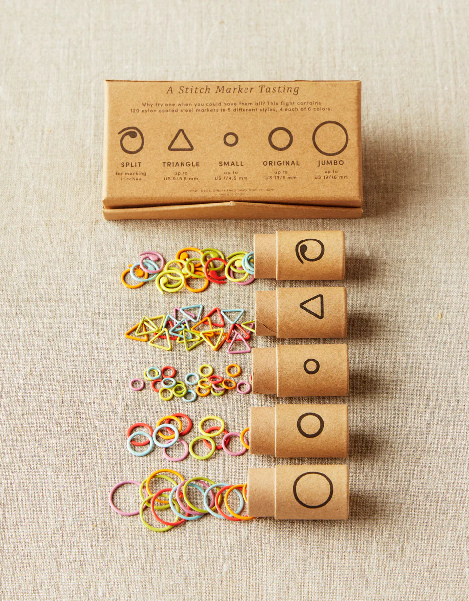 Kraft brown box of cocoknits Flight of Stitch Markers, lying on a gray surface with five open containers of colorful stitch markers. 