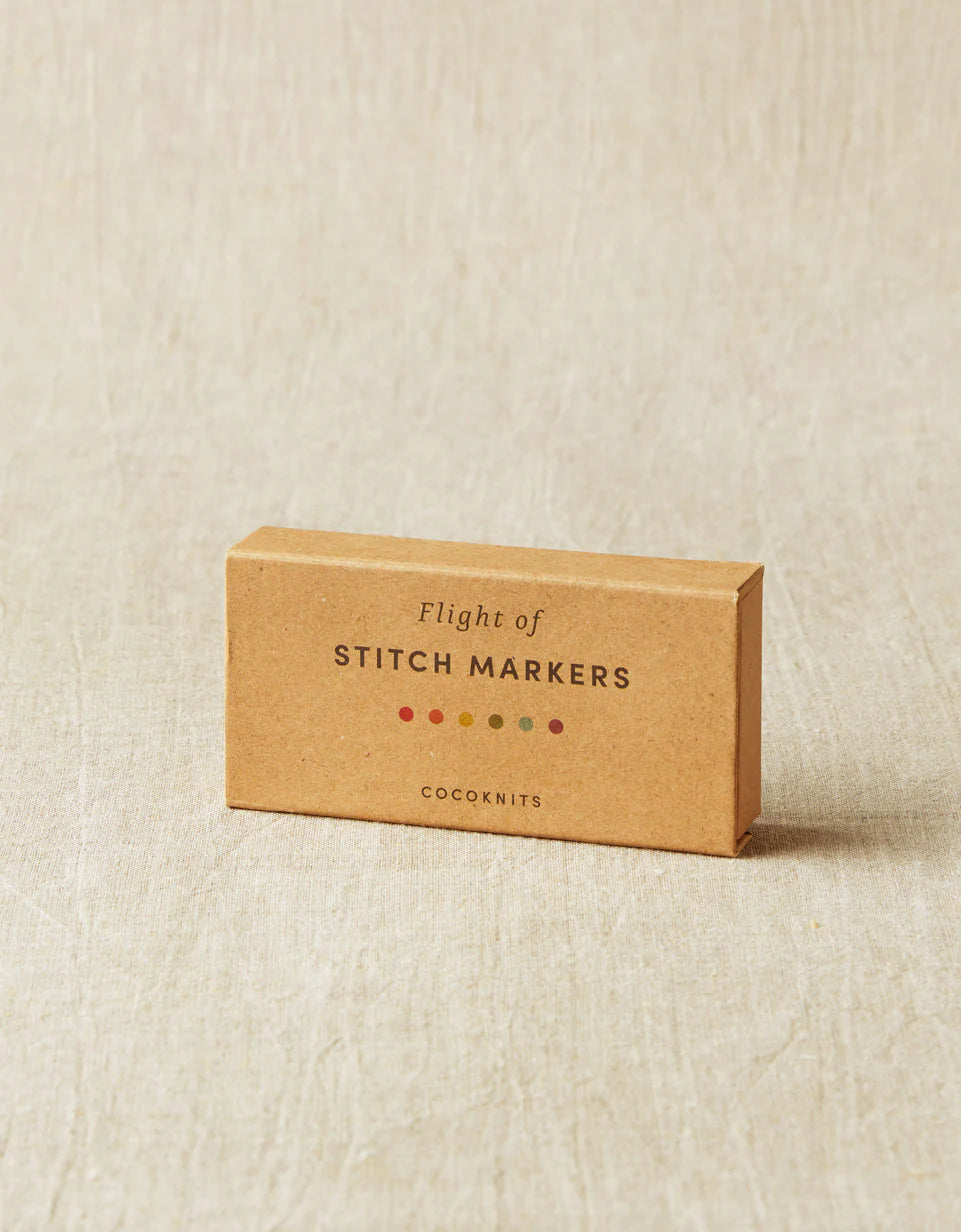 Kraft brown box of cocoknits Flight of Stitch Markers, lying on a gray surface. 