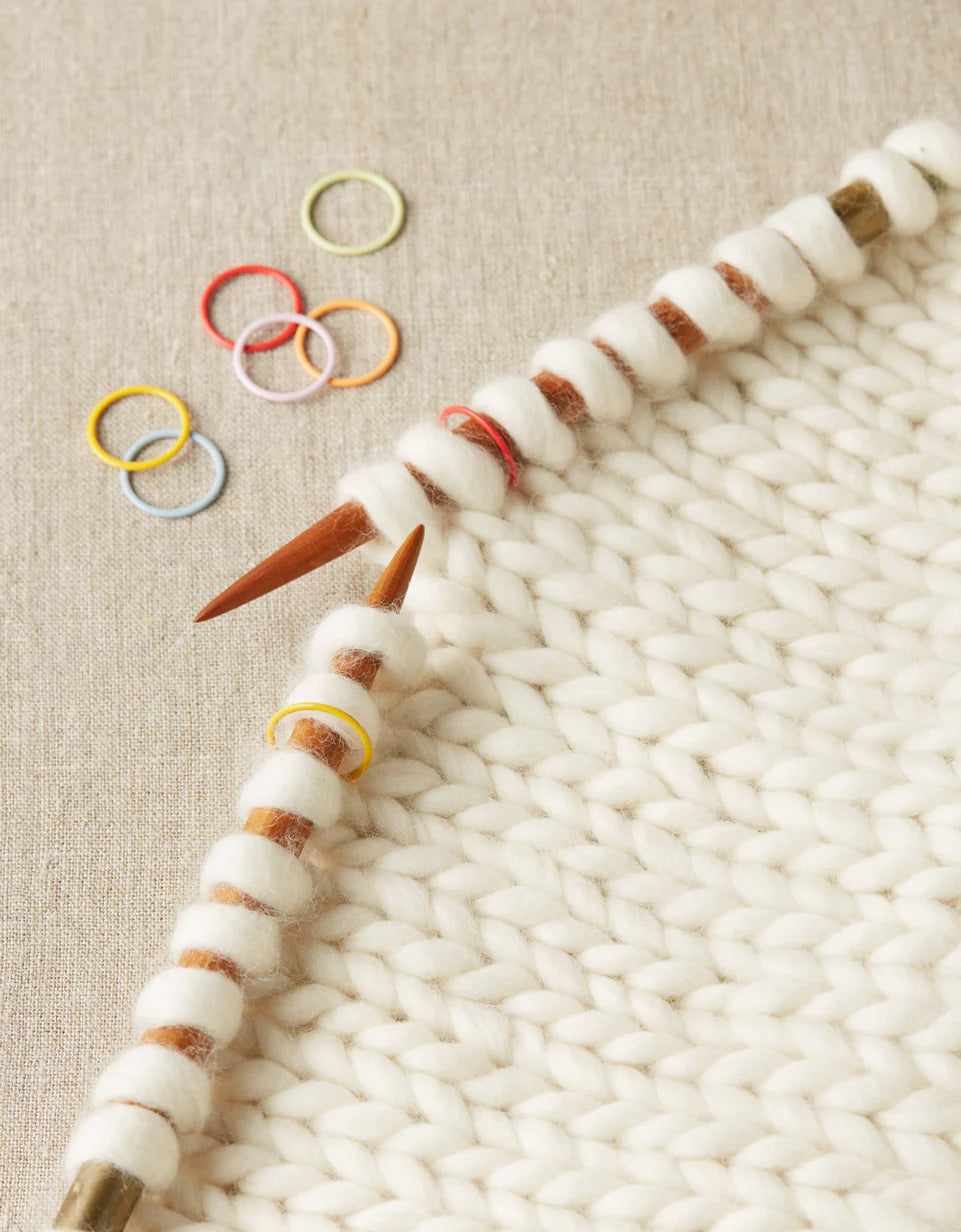 Close up on white knitting in progress, with a selection of colorful stitch markers nearby. 