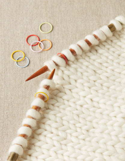 Close up on white knitting in progress, with a selection of colorful stitch markers nearby. 