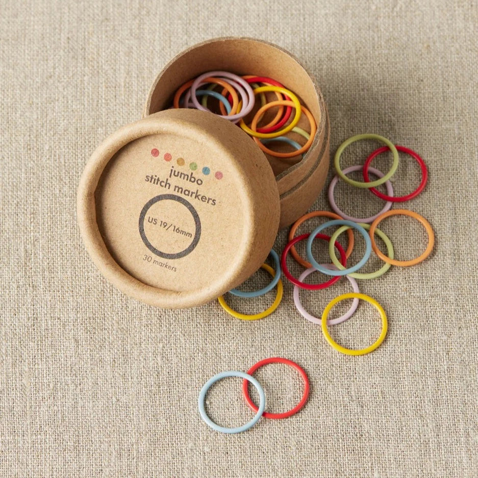 Colorful round stitch markers in pastel shades, strewn from an open box across a gray surface. 