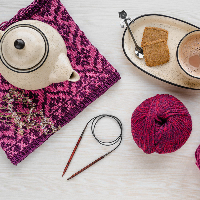 Brown circular needles connected by a black cord, laid on a table near a ball of yarn, teapot, and plate of cookies. 