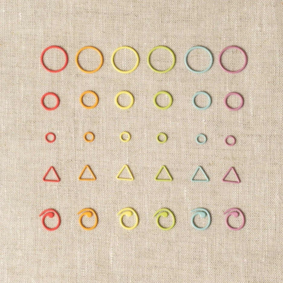 Rainbow of cocoknits stitch markers in a variety of shapes - round, triangle, curlicue - lined up on a gray surface. 