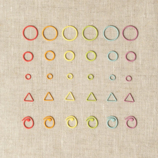 Rainbow of cocoknits stitch markers in a variety of shapes - round, triangle, curlicue - lined up on a gray surface. 