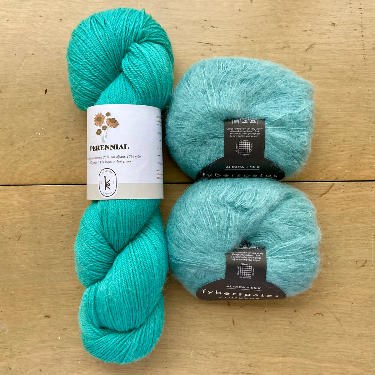 The contents of an "Oslo Hat" Kit - Hillsborough Yarn Shop edition are laid out on a wooden surface: one skein of Kelbourne Woolens Perennial in Caribbean and two balls of Fyberspates Cumulus in Sea Green. 