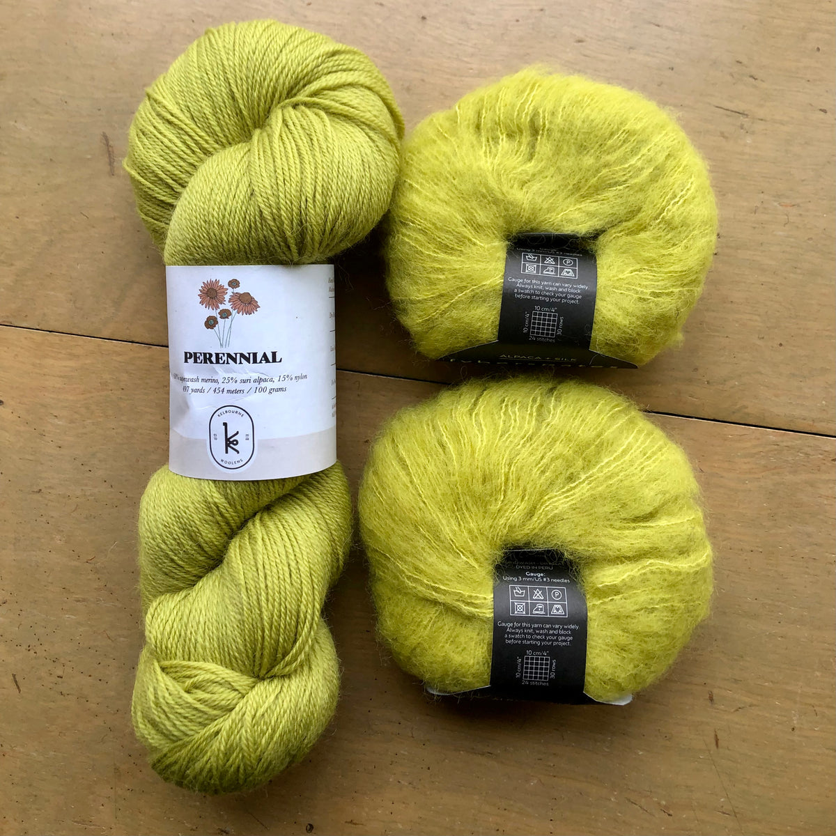 The contents of an "Oslo Hat" Kit - Hillsborough Yarn Shop edition are laid out on a wooden surface: one skein of Kelbourne Woolens Perennial in Chartreuse and two balls of Fyberspates Cumulus in Sphagnales. 